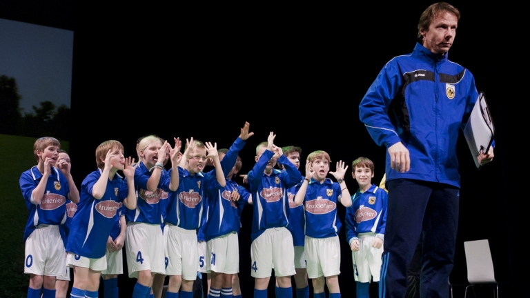 Wel winnen, hè!: coach Bas in een trainingsjack voor een juichend jeugdteam in blauwe shirts
